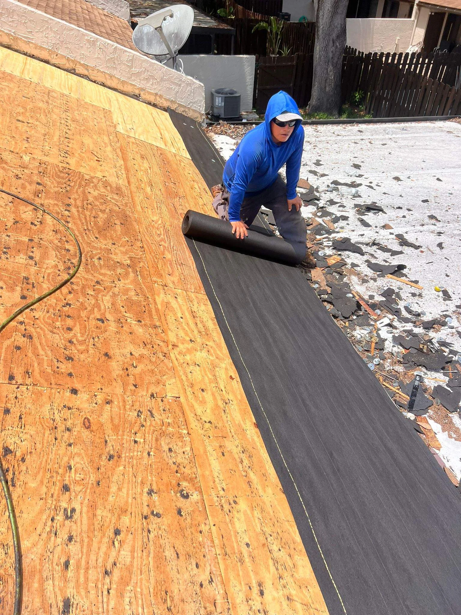 Worker installing black roofing membrane on residential roof with exposed wood sheathing.
